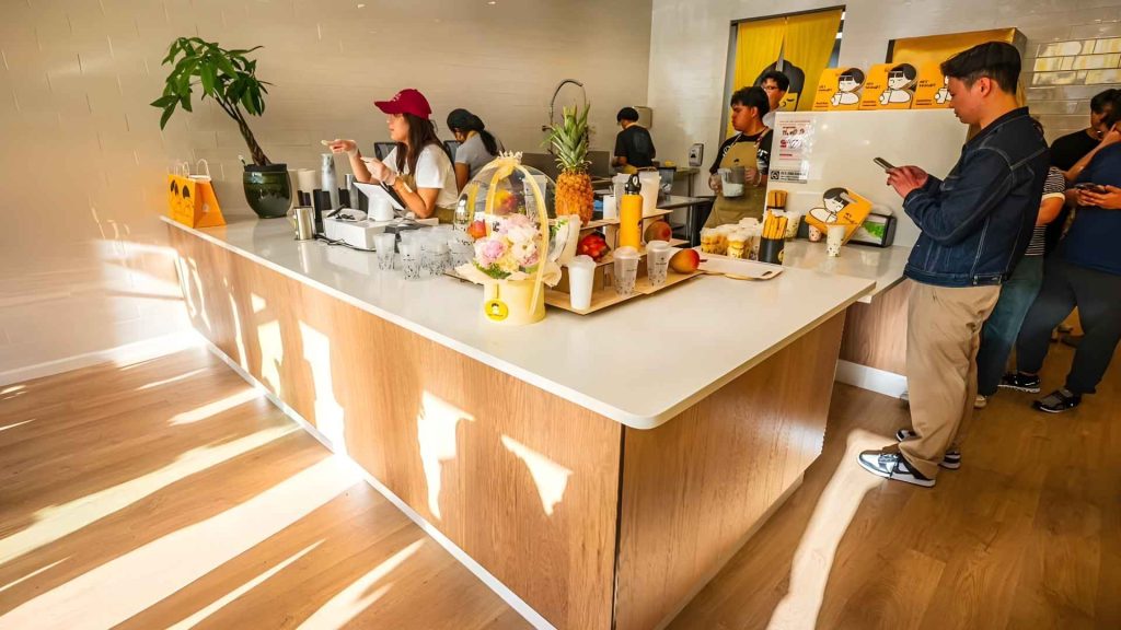 A bright and modern café scene with customers waiting at a counter where staff prepare drinks, surrounded by fresh fruit, drinks, and decorative items on a clean white countertop.