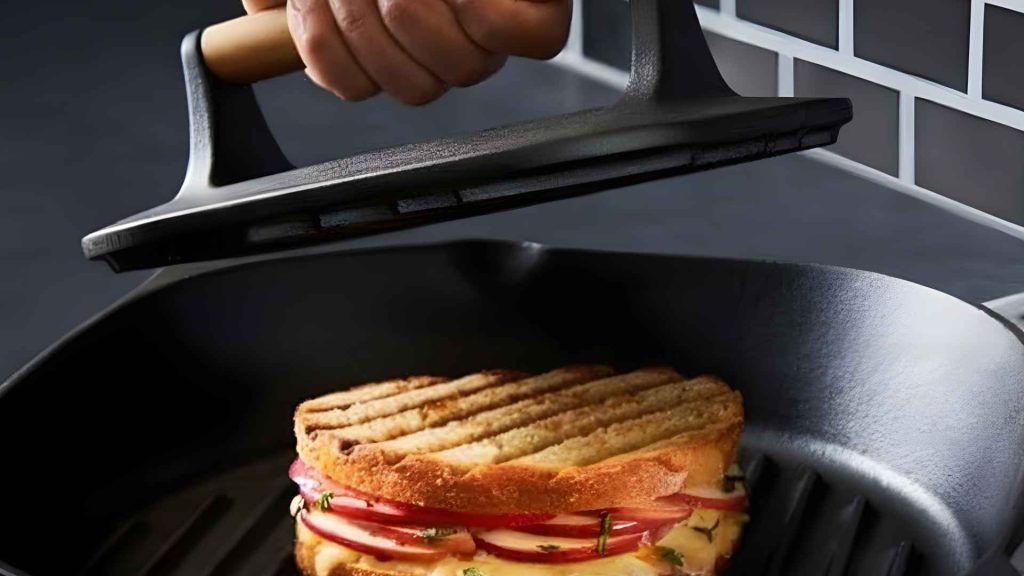 Cast iron grill press flattening a burger patty on a grill.