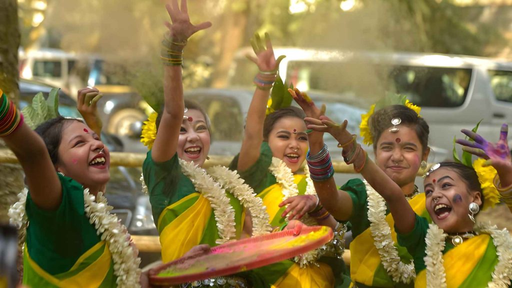 Young girls in green and yellow sarees dancing and throwing colors during Pahela Falgun celebrations.