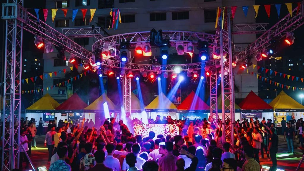 A vibrant crowd dancing under neon lights and colorful bunting at the Rishka Festival 3 outdoor music stage.
