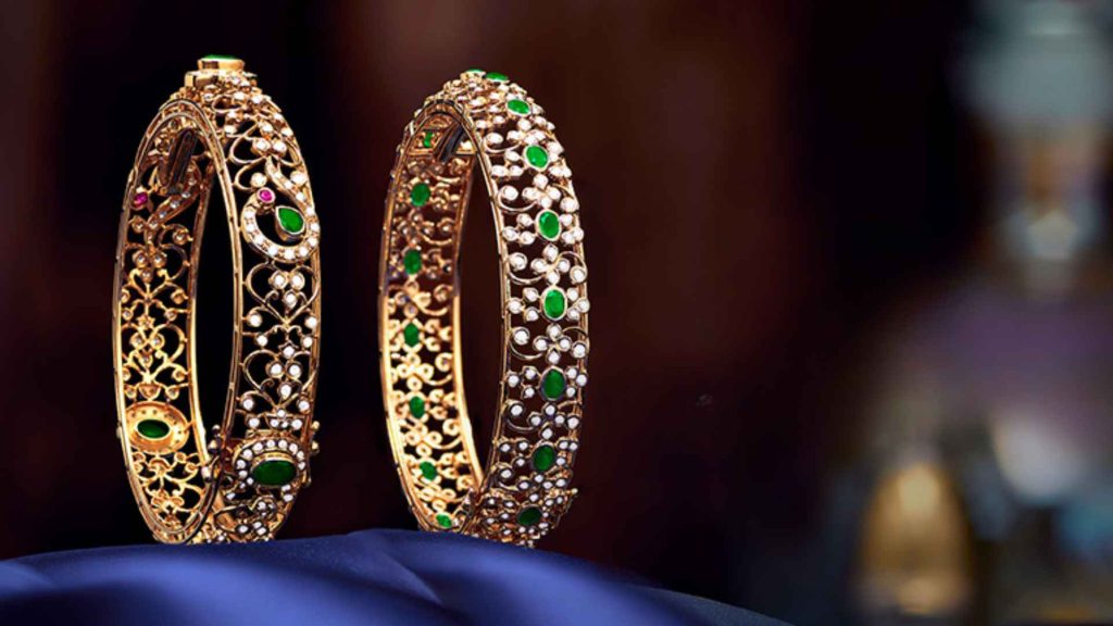 A pair of intricate gold filigree bangles set with green emeralds and diamonds by Malabar Gold & Diamonds.