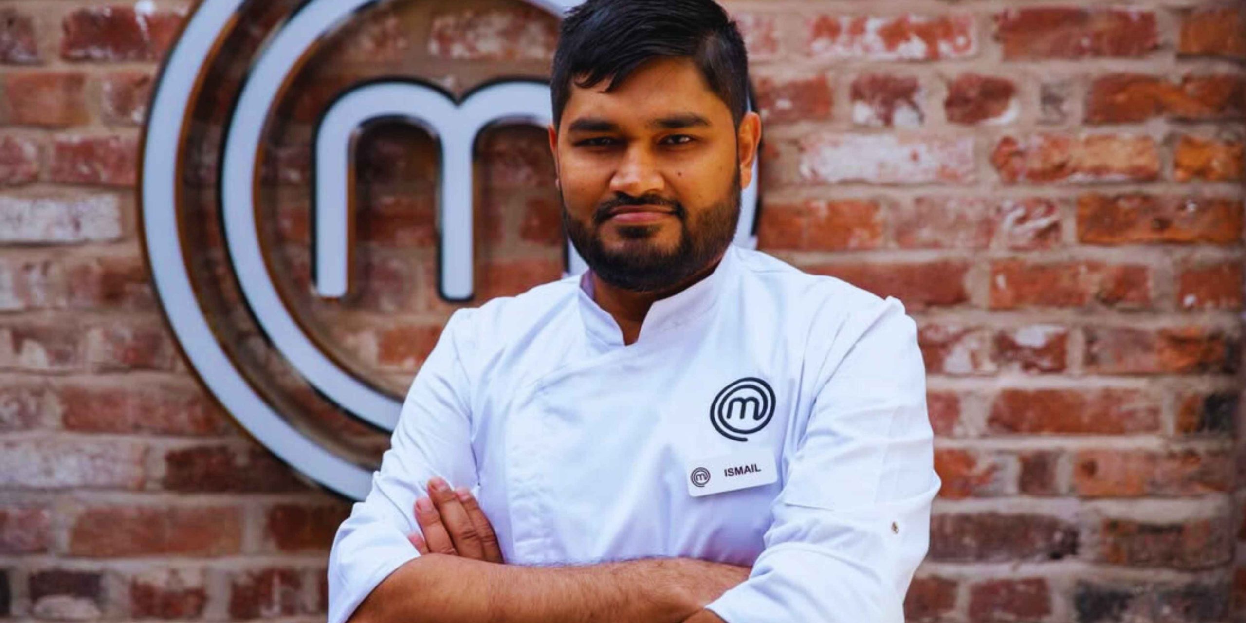 A portrait of Chef Ismail wearing a white professional chef’s coat with the MasterChef logo, standing confidently against a red brick wall during a MasterChef Historic Win.