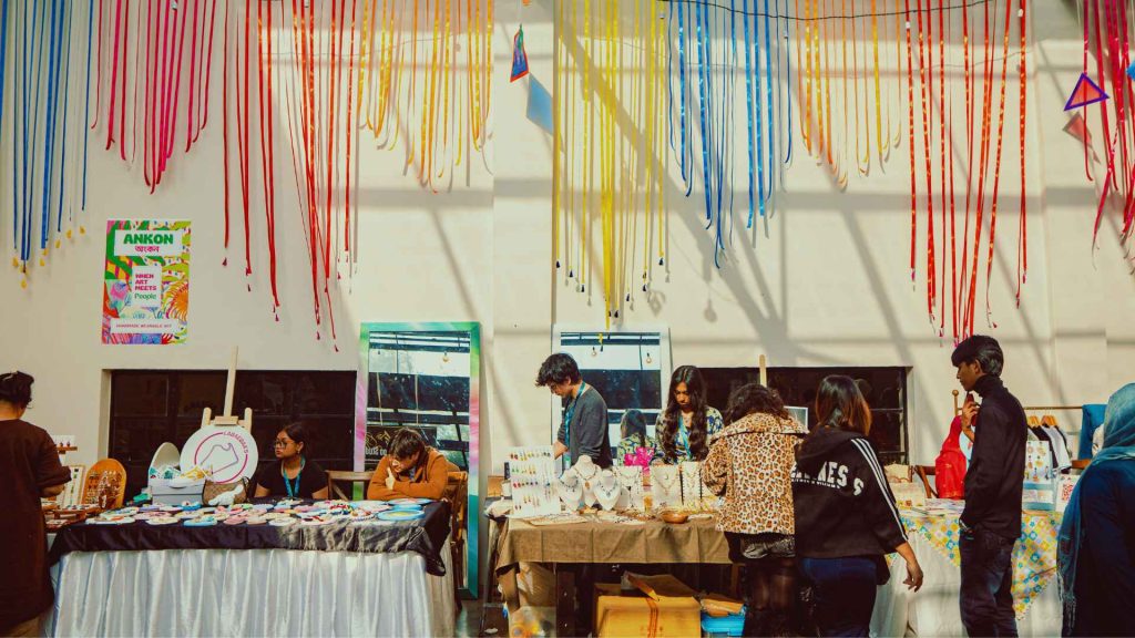 A bustling indoor art exhibition and creative market space for the Arka Boishakh 1433 festival. Tables are filled with art prints and handmade wearable art, with young attendees browsing the creative displays under colorful hanging streamers.