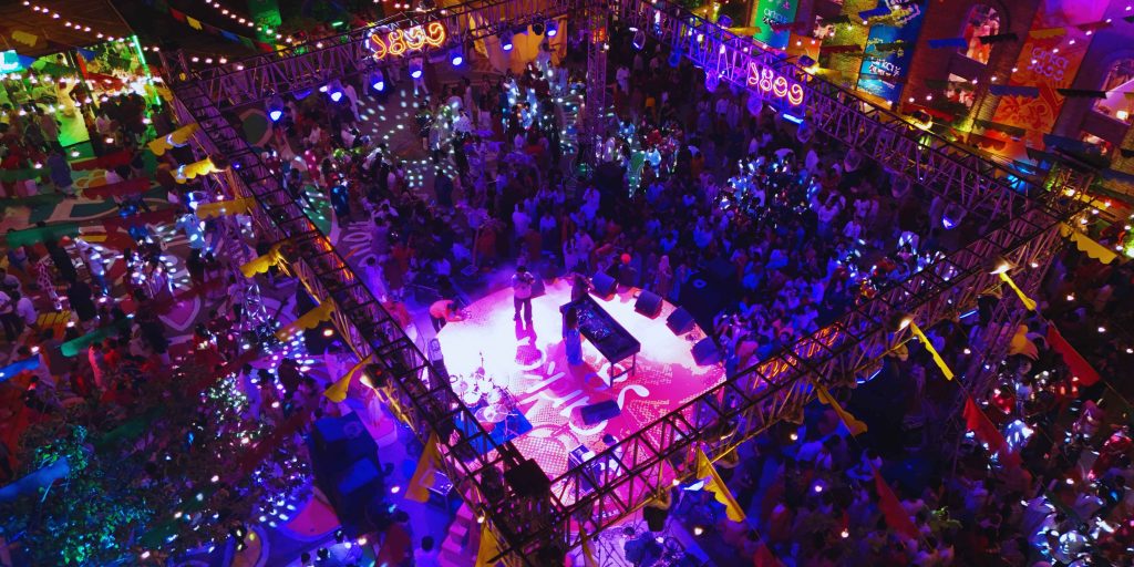 An aerial nighttime shot of a massive, energetic crowd celebrating under glowing neon truss structures and colorful fusion lighting at the Arka Boishakh 1433 festival in Dhaka.