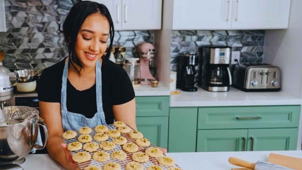 Shompa Kabir MasterChef smiling in her home kitchen while holding a wire rack of freshly baked pistachio-topped cookies.