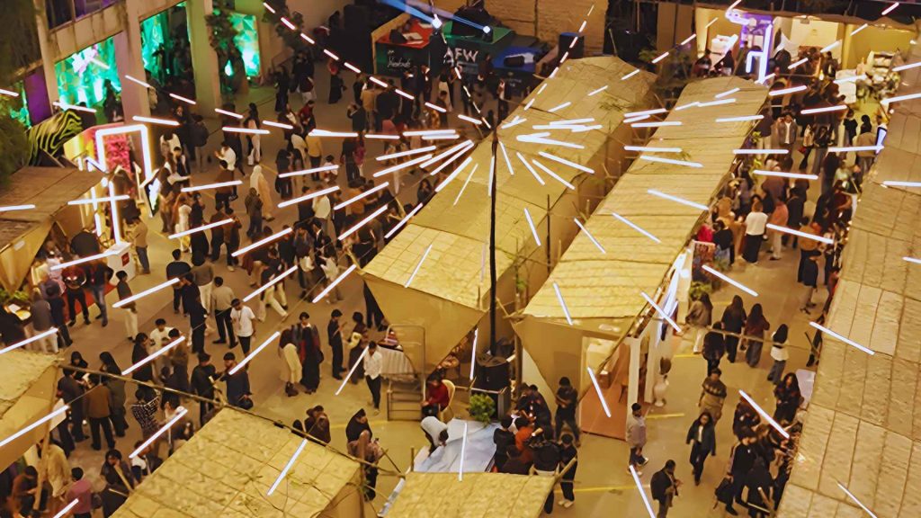 High-angle night photograph of the bustling and illuminated Arka Boishakh 1433 festival grounds in Dhaka, showing crowds of people browsing thatched-roof stalls under unique geometric light installations.