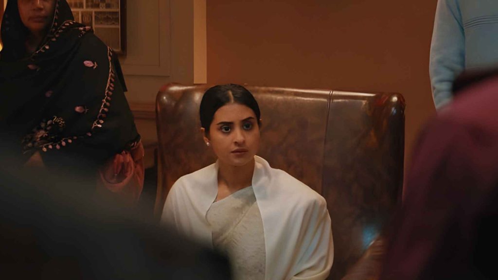 A dramatic scene from the film Pressure Cooker showing a worried woman in a white saree sitting in a leather chair, surrounded by people.