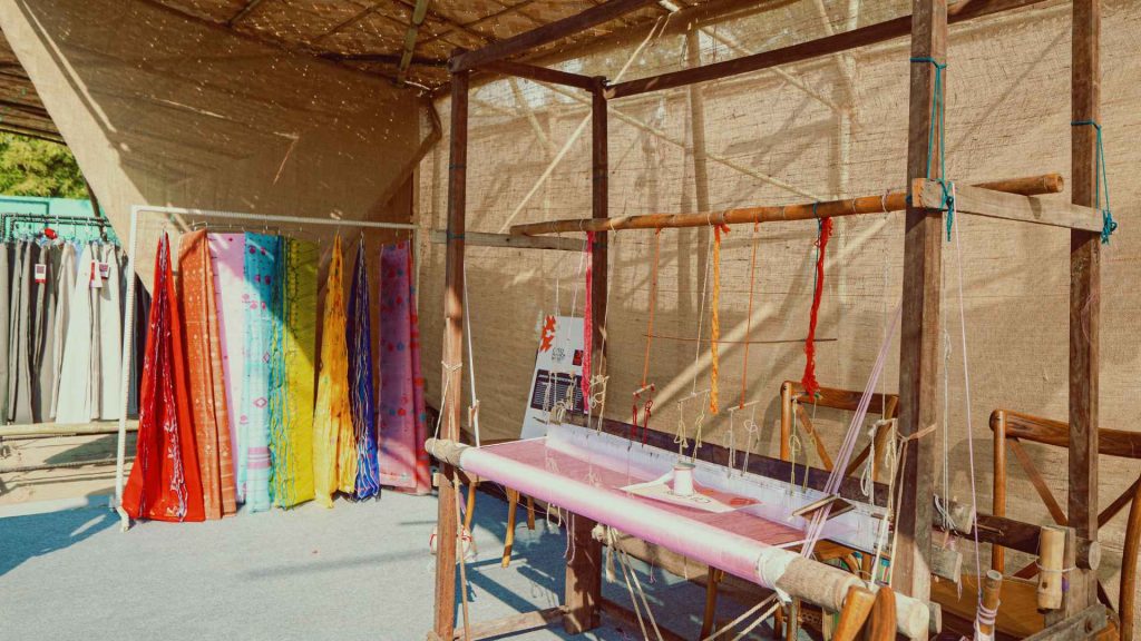 A traditional wooden handloom with pink yarn is set up under a jute-covered rustic structure. In the background, colorful, rainbow-patterned Bengali sarees hang on a rack, showcasing textile art during the Arka Boishakh 1433 festival.