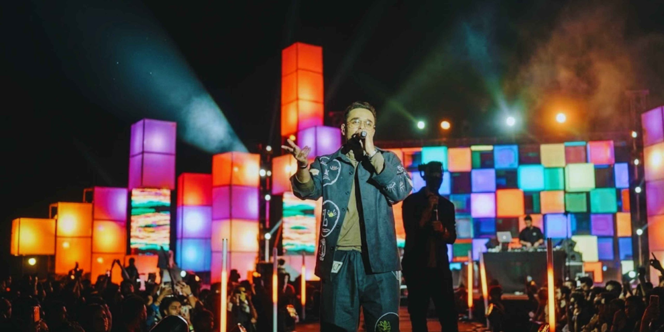 Alemam Elghandour performing live on a grand outdoor stage featuring vibrant, multi-colored cube lighting and a massive crowd during a music festival.
