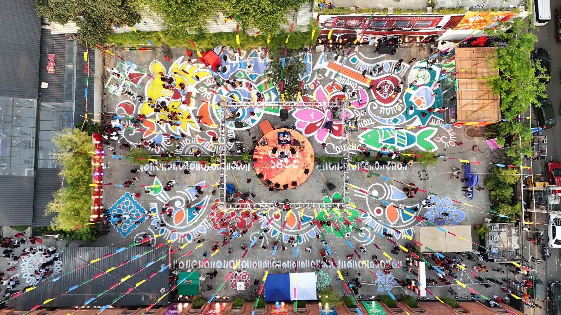 An aerial drone photograph of the vibrant outdoor venue Aloki in Dhaka, displaying massive colorful alpona, the central stage, and thousands of attendees celebrating Arka Boishakh 1433