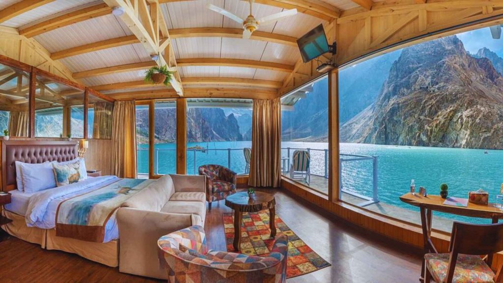 A luxury bedroom suite at Luxus Hunza with a king-sized bed, seating area, and a massive glass wall showcasing the turquoise Attabad Lake and mountain peaks.