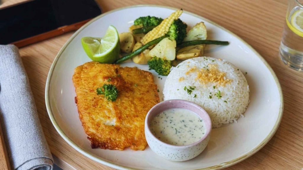 Close-up of a plated main course featuring golden fried fish, a mound of garlic rice with garnish, a bowl of dipping sauce, and grilled vegetables at Zen Café. Ranked highly on the Best Café in Dhanmondi menu.