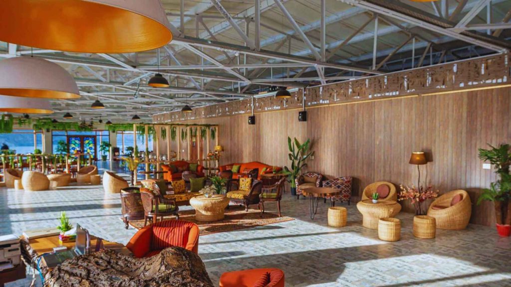 The expansive indoor restaurant and lounge at Luxus Hunza featuring high-industrial ceilings, elegant wicker seating, and large windows overlooking Attabad Lake.